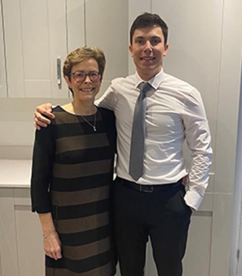 Mother in a black dress stood next to her son in a white shirt, blue tie and black trousers. He has his arm around her. 