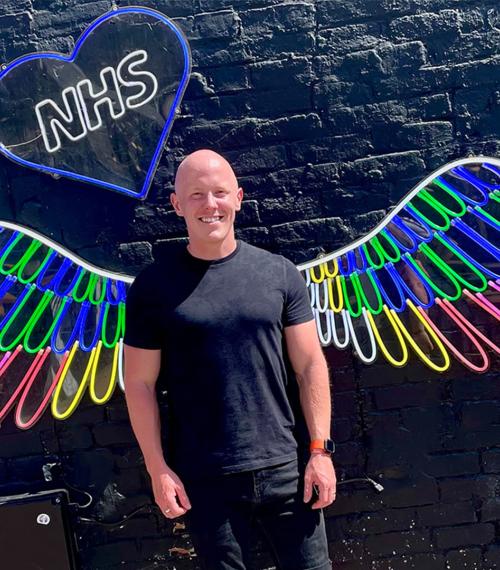 Person stood in front of a black wall with neon wings and NHS in a blue heart. 