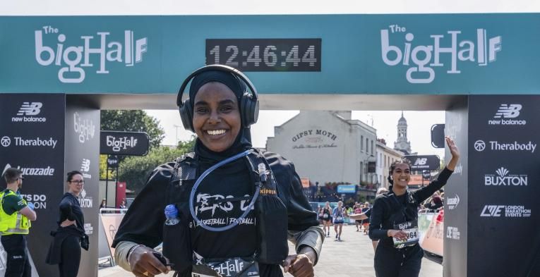 Smiling woman wearing a rucksack and headphones crosses the finish line. woman running behind her waves to the spectators.