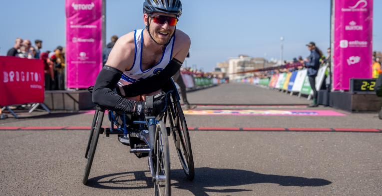 Man in racing chair poses at the finish line of Brighton Marathon.