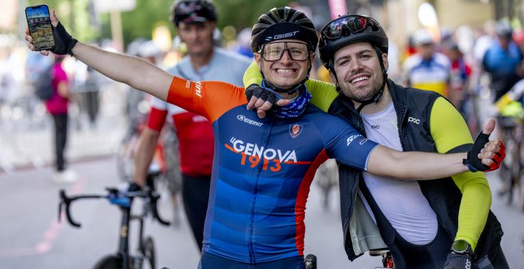 Two cyclists pose together, one has his arms outstretched and the other has his arm around the first cyclist.