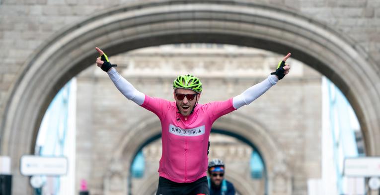 Cyclist on a bike points both hands up smiling as he rides over London bridge.