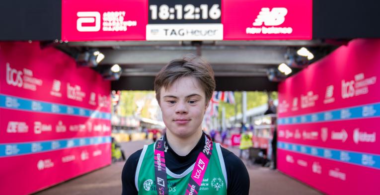 Young person stood in front on the London Marathon finish line. With a medal around their neck. 