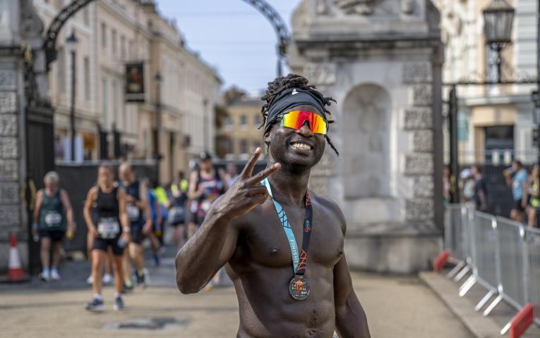 Man stands topless wearing a medal and sunglasses making the peace sign.