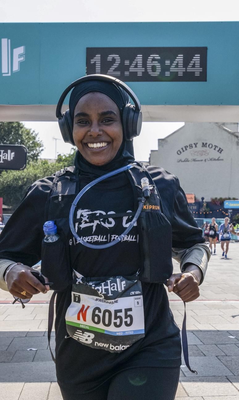 Smiling woman wearing a rucksack and headphones crosses the finish line. woman running behind her waves to the spectators.