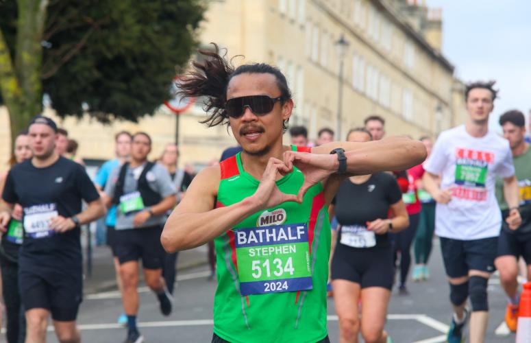 Person running in a green vest with sunglasses on making a heart sign with their fingers