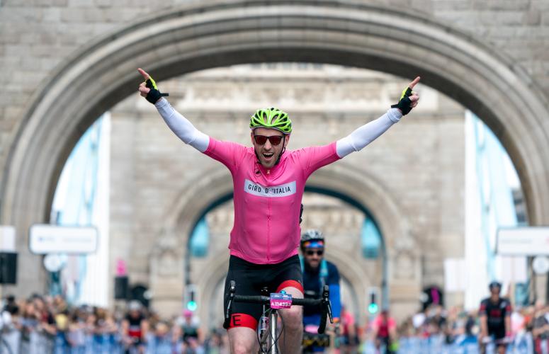 Cyclist on a bike points both hands up smiling as he rides over London bridge.