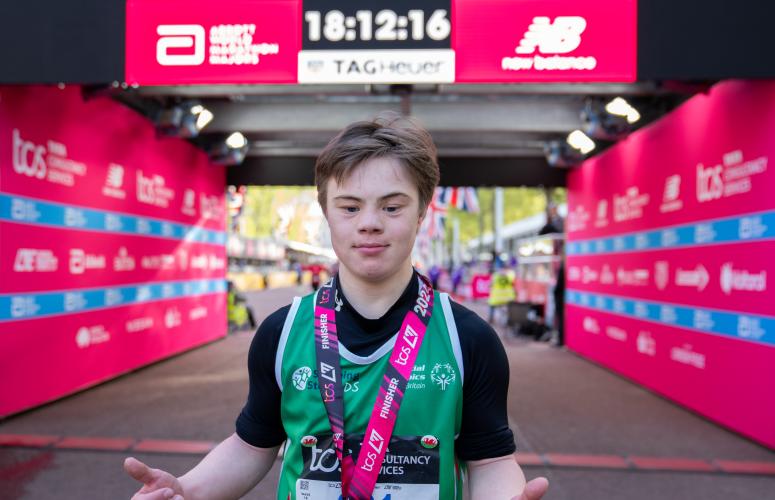 Young person stood in front on the London Marathon finish line. With a medal around their neck. 