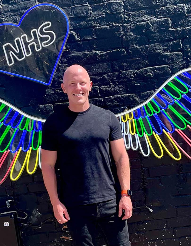 Person stood in front of a black wall with neon wings and NHS in a blue heart. 