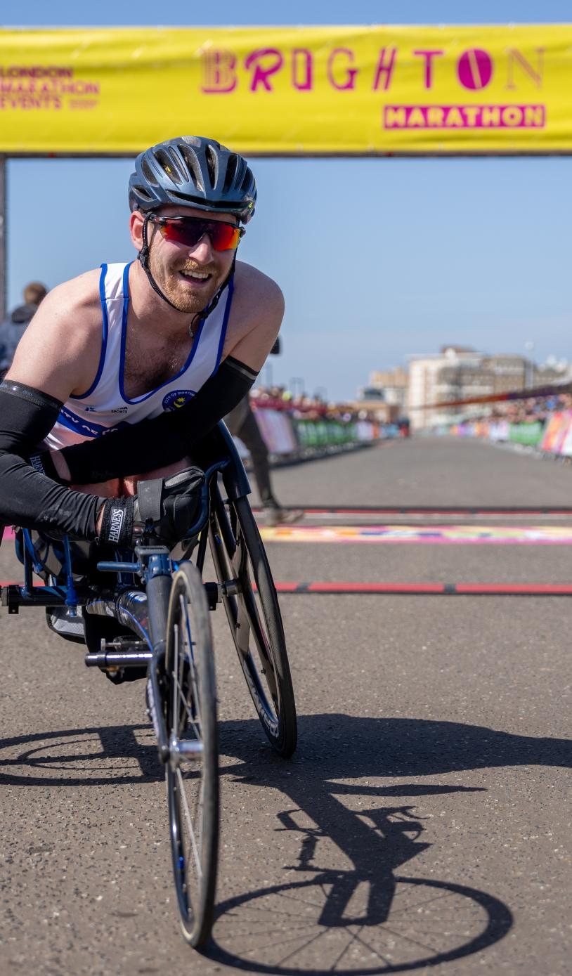 Man in racing chair poses at the finish line of Brighton Marathon.