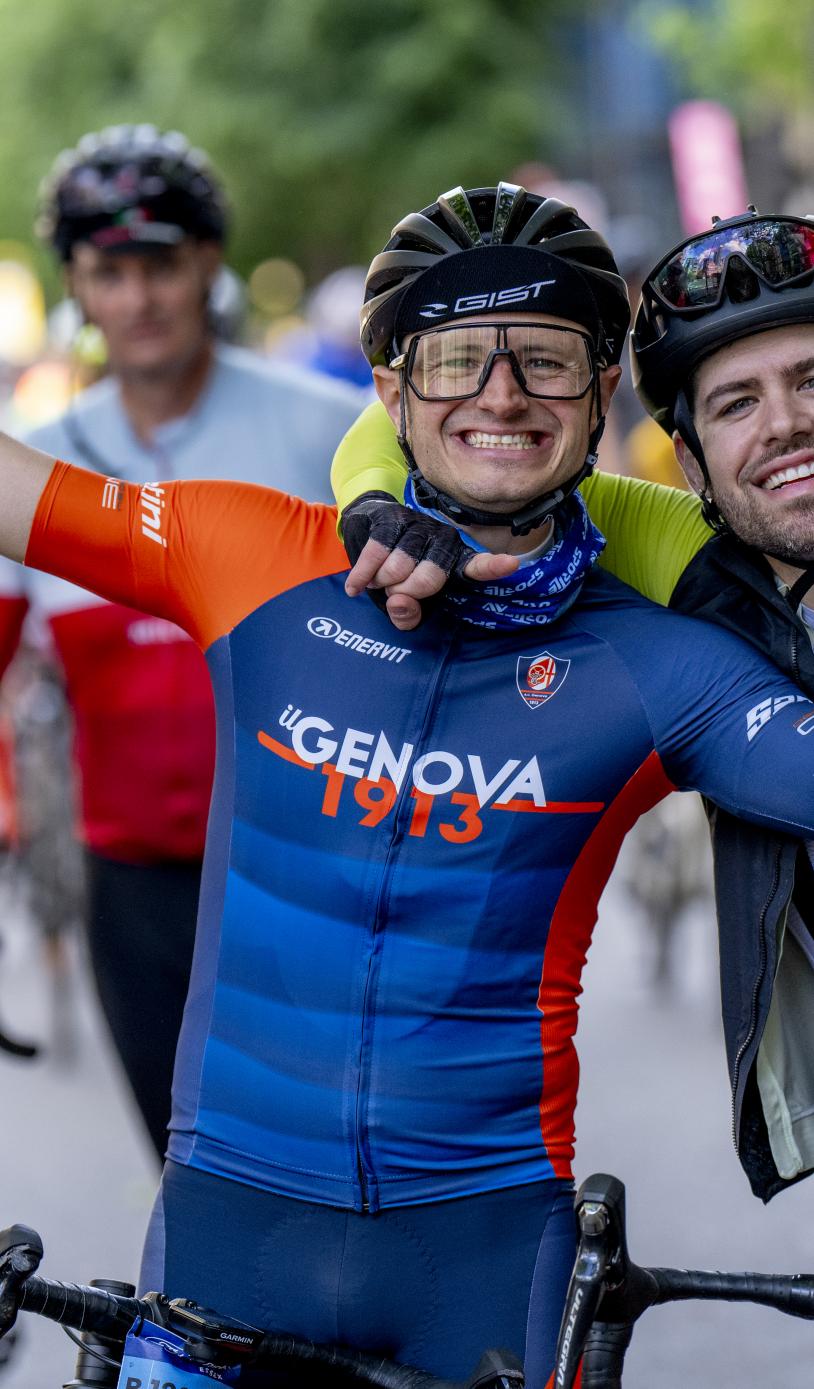 Two cyclists pose together, one has his arms outstretched and the other has his arm around the first cyclist.