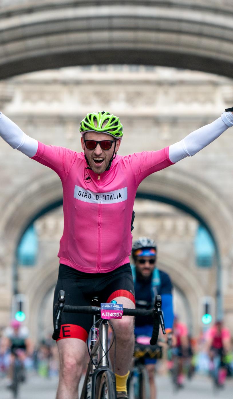 Cyclist on a bike points both hands up smiling as he rides over London bridge.