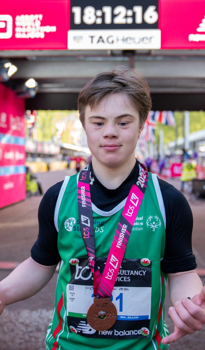 Young person stood in front on the London Marathon finish line. With a medal around their neck. 