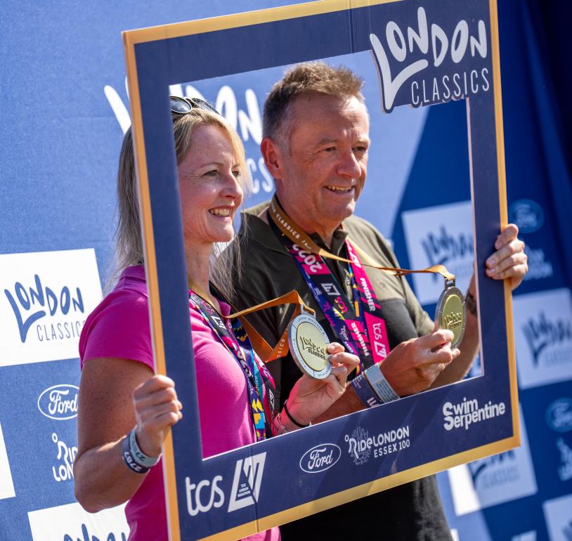 Two people holding up their London Classics medals and a cardboard frame in front of them