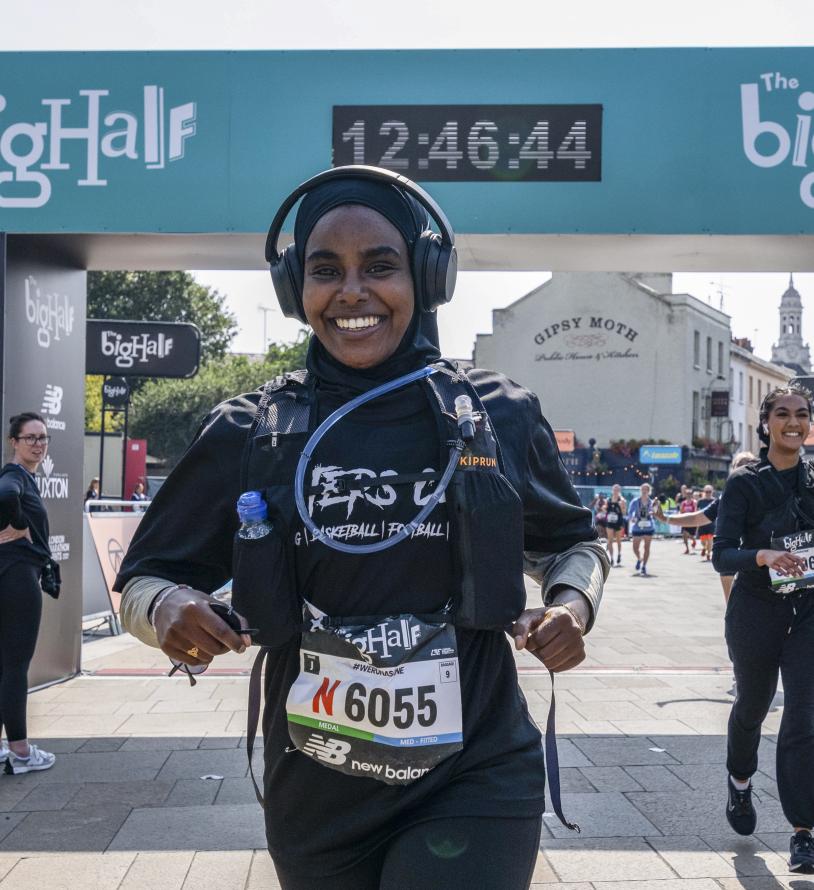 Smiling woman wearing a rucksack and headphones crosses the finish line. woman running behind her waves to the spectators.